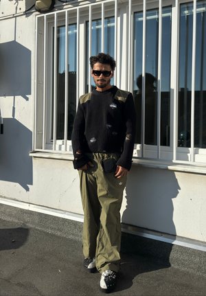 Suéter negro de canalé con detalles desgastados y acentos en verde oliva en los hombros, combinado con pantalones sueltos en oliva y zuecos negros decorativos.