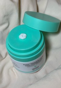 Ein türkisfarbener Krug mit passendem Deckel, der ein weißes Etikett mit Text trägt. Der Deckel ist leicht geöffnet und gibt eine cremige Substanz im Inneren preis.