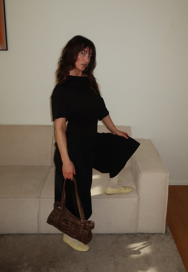 Naine mustas kleidis ja koorevärvi kingades poseerib ühe põlvega beežil diivanil, hoides pruuni käekotti tavalise valge seinaga taustal.