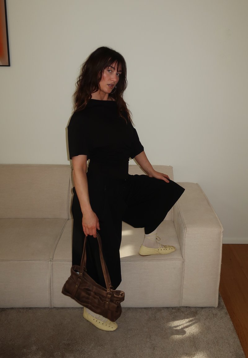 Femme en robe noire et chaussures crème posant avec un genou sur un sofa beige, tenant un sac à main marron contre un mur blanc uni.