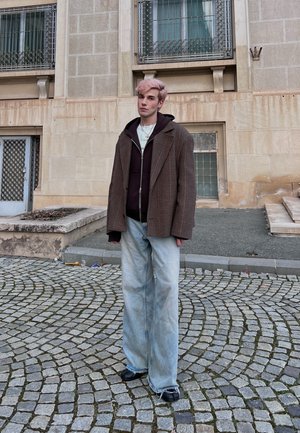 Joven con cabello rosa de pie en una calle adoquinada, vistiendo un blazer a cuadros grande, sudadera negra con capucha, jeans anchos claros y zapatos negros.