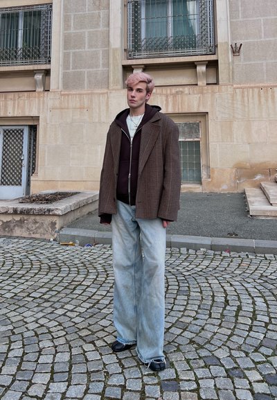 Joven con cabello rosa de pie en una calle adoquinada, vistiendo un blazer a cuadros grande, sudadera negra con capucha, jeans anchos claros y zapatos negros.