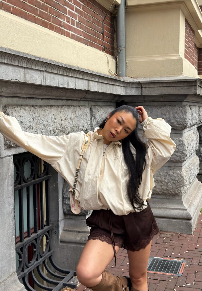 Jeune femme aux longs cheveux appuyée contre un mur en pierre, portant une veste crème ample, un short en dentelle foncé et des bottes beige montant jusqu’aux genoux sur le trottoir en ville.