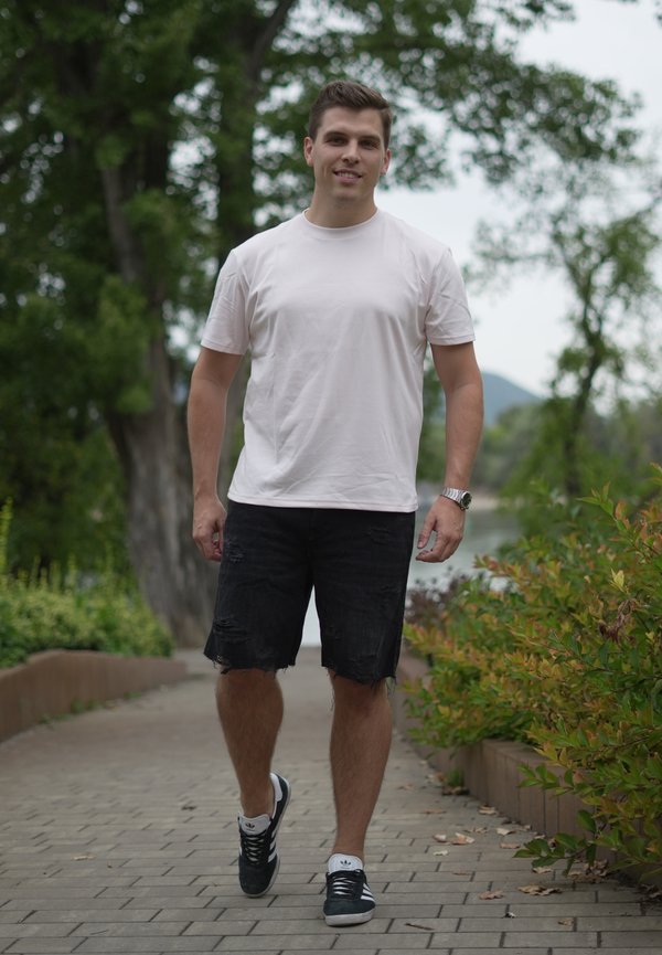 Ung mand i hvid t-shirt, sorte revnede shorts og sneakers går på en brolagt sti omgivet af grøn natur udendørs.