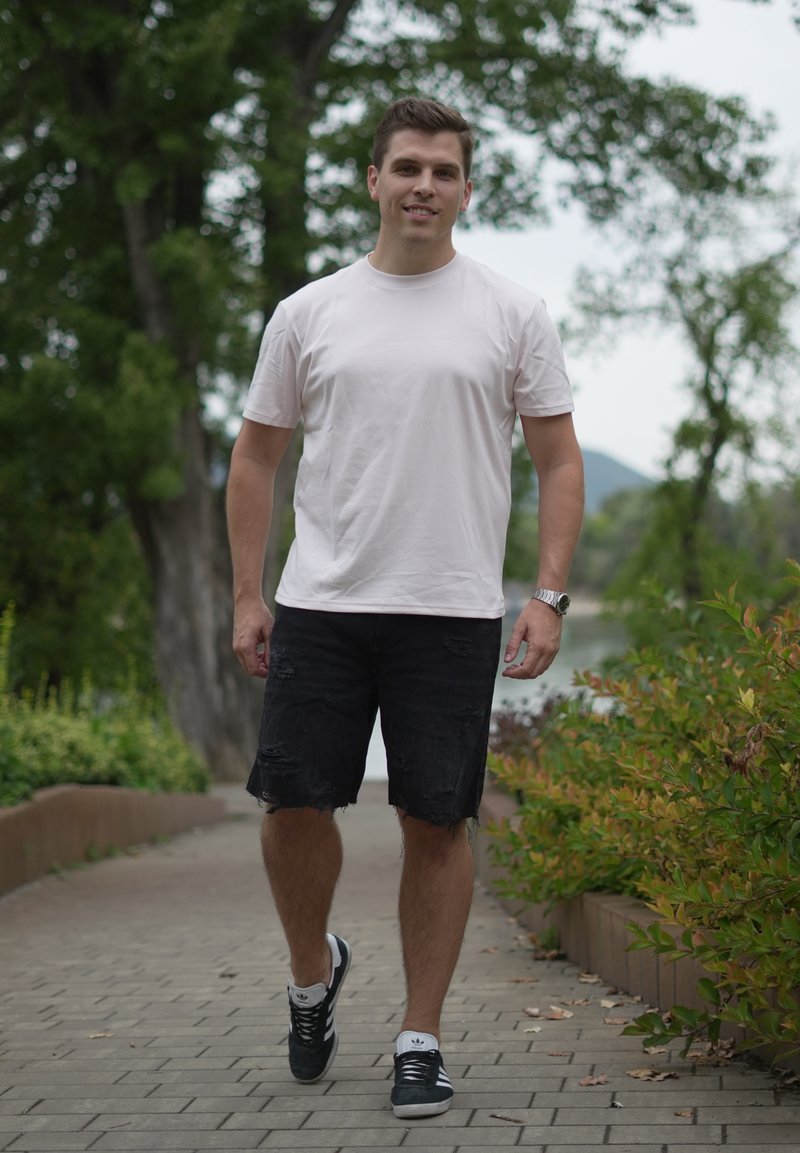 Jeune homme en t-shirt blanc, short noir déchiré et baskets marchant sur un chemin pavé entouré de verdure à l'extérieur.