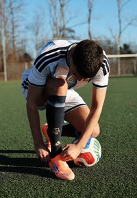 Joueur de football à genoux sur le gazon artificiel, attachant ses chaussures à crampons orange près d'un ballon de football multicolore, portant un uniforme blanc avec des rayures noires.
