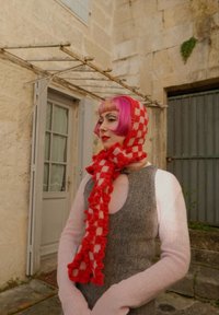 Rød og hvit rutete strikket skjerf med frynser, brukt med en grå ermeløs kjole og rosa langermet skjorte i et steinmiljø.
