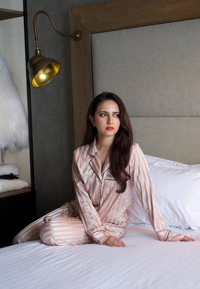 Mujer de cabello largo y oscuro, vestida con pijama de rayas rosadas y seda, sentada en una cama blanca en un dormitorio moderno con una lámpara de pared de latón.