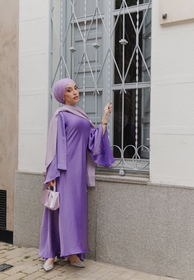 Vestido de satén púrpura con mangas anchas, combinado con una bufanda de color púrpura claro. Ella sostiene un bolso brillante de lavanda. El fondo presenta una ventana con barrotes.