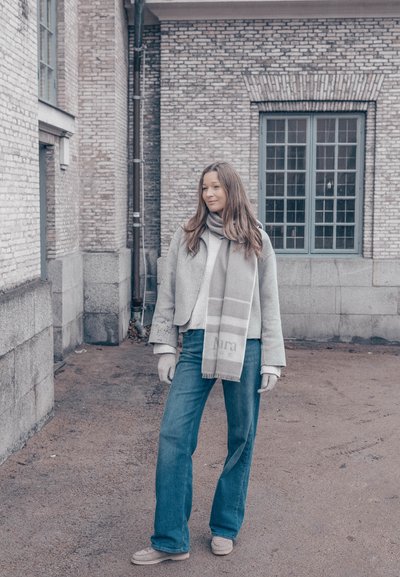 Chaqueta de lana gris, bufanda gris claro con un patrón, jeans azules y calzado beige; de pie contra una pared de ladrillo texturizada.