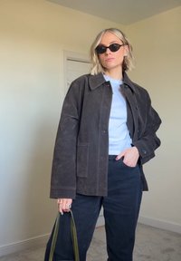 Jachetă din suede maro cu guler în formă de V, cămașă albastră deschis dedesubt și pantaloni negri. Ținând o geantă cu curea verde, purtând ochelari de soare negri.