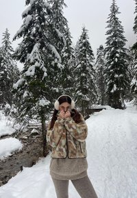 Flauschige, orange und mintgemusterte Zip-Jacke, beiger Strickpullover, hellbeige Hose und warme, weiße Ohrenschützer in einer schneebedeckten Waldlandschaft.