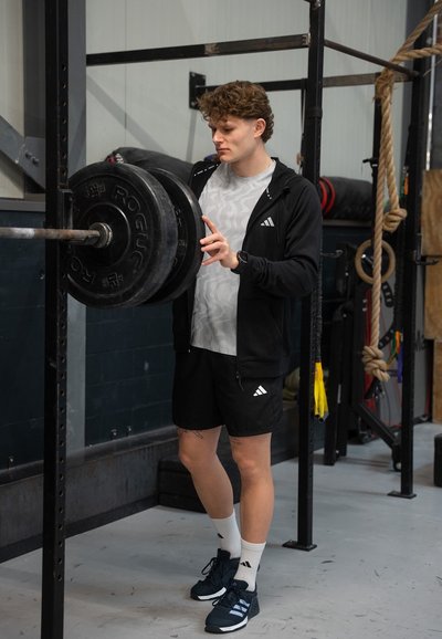 Barra de pesas con discos negros en un soporte de metal. Persona vestida con una sudadera negra con cremallera y pantalones cortos, camiseta de patrón claro y zapatillas oscuras.