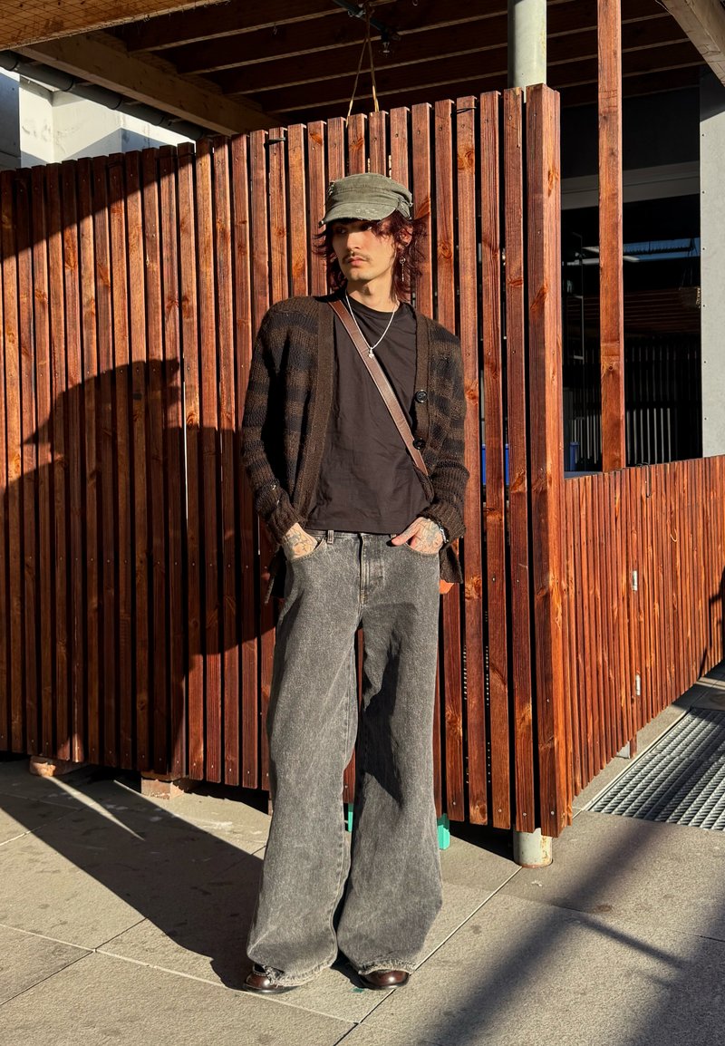 Giovane che indossa un berretto verde, un cardigan a righe scuro, una camicia nera e jeans larghi grigi, in piedi con le mani in tasca contro una recinzione di legno.