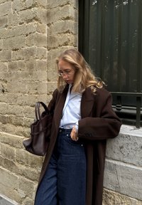 Cappotto marrone oversize su una camicia bianca, abbinato a jeans blue scuro a vita alta. Una borsa scura è tenuta di lato, con una superficie in tessuto testurizzato.