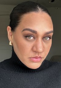 Nærbilde av et ansikt på en person iført en svart ribbestrikket turtleneck genser. Merkbare gulldråpeøredobber. Hudtonen er varm med definerte trekk.