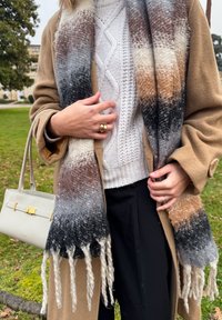 Άτομο που φοράει μπεζ παλτό, λευκό πουλόβερ πλεχτό με καλώδια, μαύρο παντελόνι και μακριά φουλάρι ομπρέ, κρατώντας ένα λευκό τσαντάκι έξω.