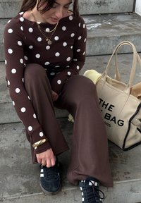 Femme assise sur des marches en béton, portant un haut et un pantalon marron à pois, en train de lacer des baskets noires, avec un sac fourre-tout beige à côté d'elle.