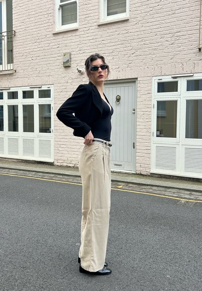 Persona con gafas de sol oscuras que lleva un top negro y una blazer, pantalones beige de pierna ancha, de pie en una calle frente a un edificio de ladrillo claro.