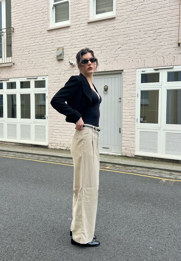 Personne avec des lunettes de soleil foncées, portant un haut noir et un blazer, un pantalon beige à jambes larges, debout dans une rue devant un bâtiment en briques clair.