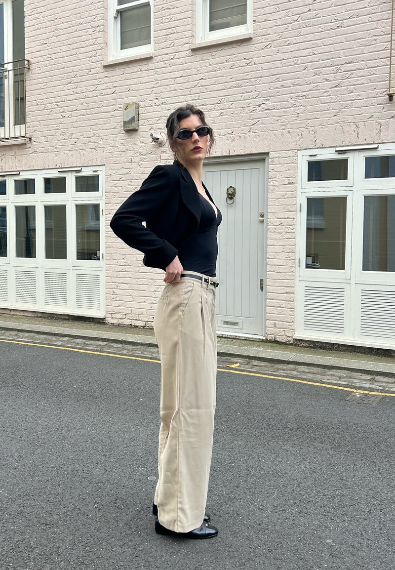 Persona con occhiali da sole scuri che indossa un top nero e blazer, pantaloni beige a gamba larga, in piedi su una strada di fronte a un edificio in mattoni chiari.