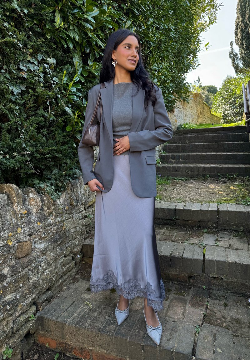 Femme debout sur des marches en pierre portant un blazer gris, une longue jupe en satin avec un ourlet en dentelle, un top gris, des talons pointus et portant un petit sac à bandoulière.