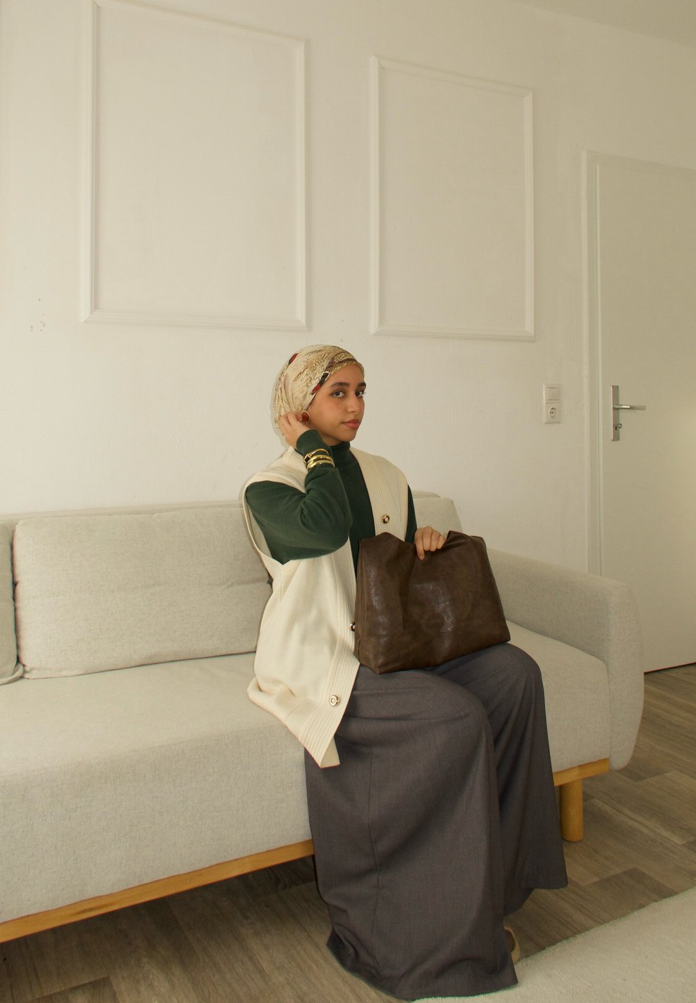 Femme portant un foulard à motifs et un gilet beige, assise sur un canapé gris clair, tenant un grand sac en cuir marron dans une pièce minimaliste.