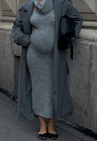 Persoana însărcinată îmbrăcată într-o rochie lungă din tricot gri, cu o haină gri supradimensionată, pantofi negri fără toc și ținând o geantă neagră, stând pe trotuarul unei oraș.