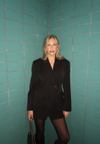 Blazer noir oversize à un bouton, porté sur une mini-jupe. En arrière-plan, des murs carrel és turquoise. Accessoire : sac à bandoulière en chaîne.