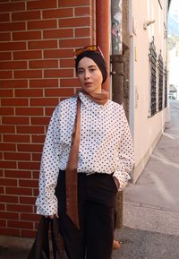 Camisa blanca con lunares negros, corbata de satén marrón, pantalones negros y un bolso marrón. La modelo lleva un pañuelo negro en la cabeza y gafas de sol.