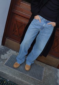 Vaaleansiniset farkut väljällä istuvuudella, yhdistettynä vaaleanruskeisiin mokkanahkakenkiin. Tumma takki näkyy osittain, taustalla puinen ovi.