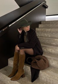 Καφέ σουέτ μπότες μέχρι το γόνατο, σκούρο παλτό, πράσινο καπέλο με φαρδύ γείσο και καφέ τσάντα. Κάθεται σε γκρι πέτρινες σκάλες με ελάχιστο φόντο.