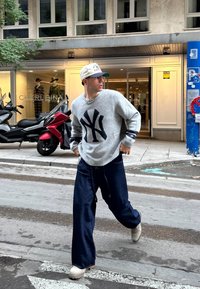 Grijze trui met marineblauw logo, losse donkere denim broek en beige schoenen. Draagt een lichtgekleurde pet en een ketting, lopend op straat.