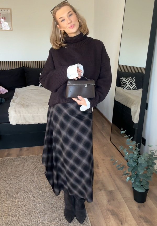 Jeune femme en pull sombre et jupe à carreaux tenant un petit sac à main noir, debout dans une pièce moderne avec un lit et un grand miroir.