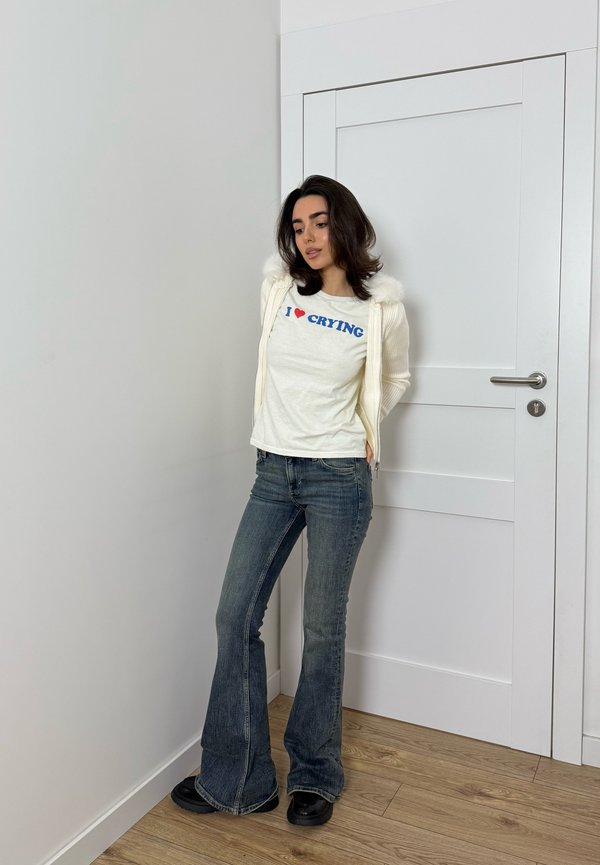 Cremefarbene Zip-Up-Jacke mit Kunstpelzdetails, weißes T-Shirt mit blauem Text "I ♥ CRYING", blaue Schlagjeans, schwarze Schuhe, Hintergrund aus Holzfußboden.