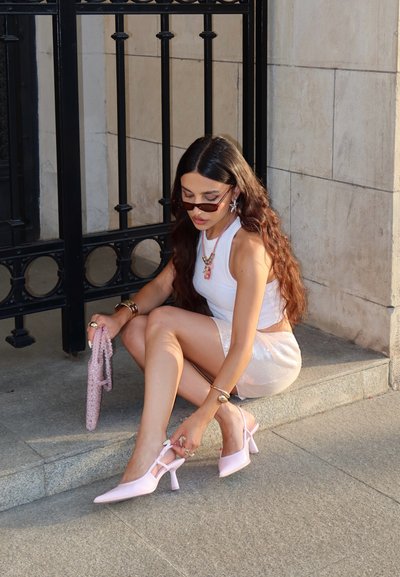 Tacones slingback de color rosa claro con punta afilada y tacón de bloque, usados por una persona sentada en un escalón de piedra, sosteniendo un bolso de cuentas pink.