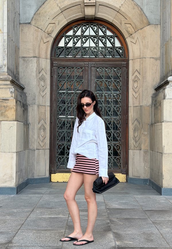 Femme portant une chemise blanche à manches longues, une mini-jupe rayée rose et noire, des lunettes de soleil noires et des sandales, tenant une pochette noire devant une porte en arc ornée.