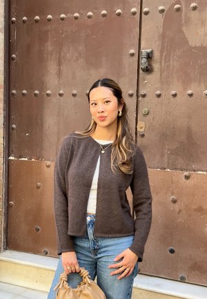 Cárdigan de lana marrón con diseño corto, llevado sobre una camisa blanca, combinado con jeans de cintura alta y un bolso marrón claro. Joyas simples de plata.