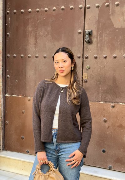 Cárdigan de lana marrón con diseño corto, llevado sobre una camisa blanca, combinado con jeans de cintura alta y un bolso marrón claro. Joyas simples de plata.