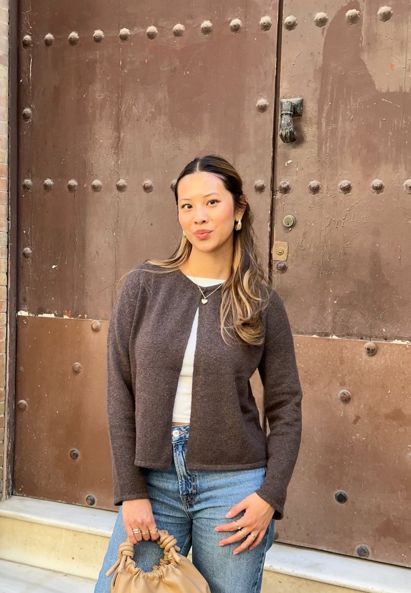 Cardigan en laine marron au design court, porté par-dessus une chemise blanche, associé à un jean taille haute et un sac à main beige. Bijoux argentés simples.