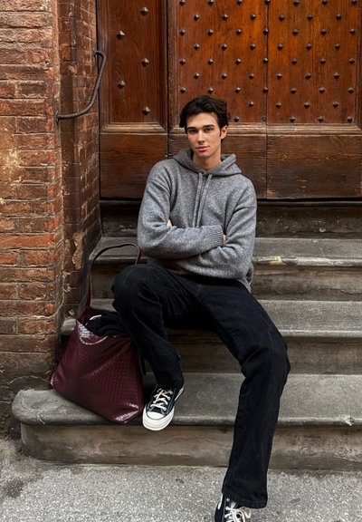 Sudadera de punto gris, jeans negros, zapatillas azul marino y un bolso trenzado burdeos. Puerta de madera característica con clavos decorativos en el fondo.