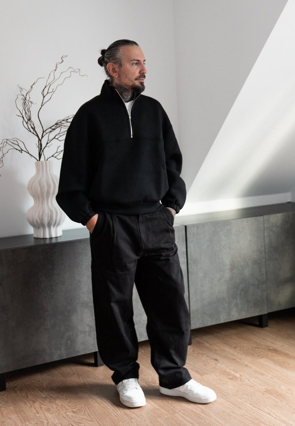 Un homme barbu se tient debout, les mains dans les poches. Il porte un haut noir à demi-zip, un pantalon noir ample et des baskets blanches dans une pièce moderne.