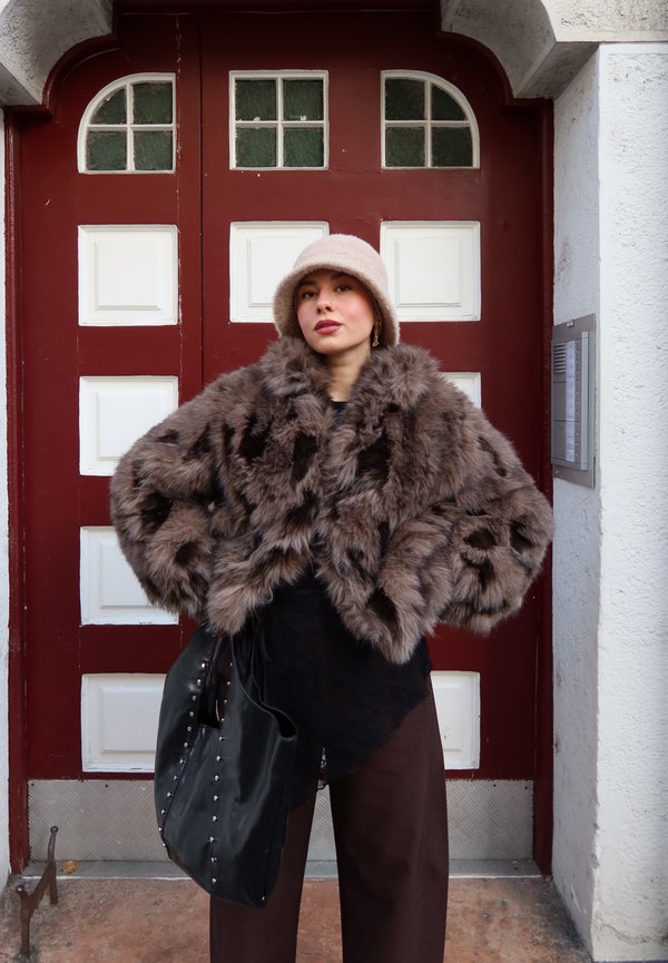 Kvinde iført beige hat, brun imiteret pelsjakke, mørke bukser, bærende en sort nittebesat taske, stående foran en dør med røde og hvide paneler.