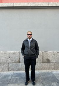 Jaqueta bomber de couro preto com fecho, camisa branca e gravata, calças pretas, óculos escuros escuros, em pé contra uma parede cinza com base de pedra.
