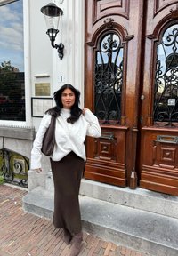 Pārmērīgi liels balts adīts džemperis un brūna svārku garums līdz elkonim; brūni papēžu zābaki; stāvot pie izsmalcinātiem koka durvīm ar dekoratīvu metāla apdari.