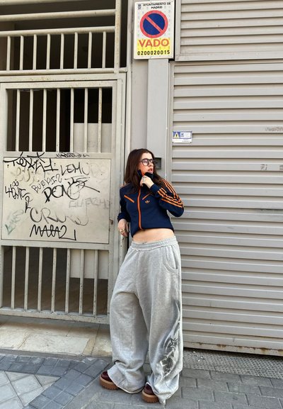 Chaqueta azul y naranja con cremallera, pantalones de chándal grises oversize con gráfico lateral, zapatos de plataforma marrones. Entorno urbano con graffiti y un cartel.