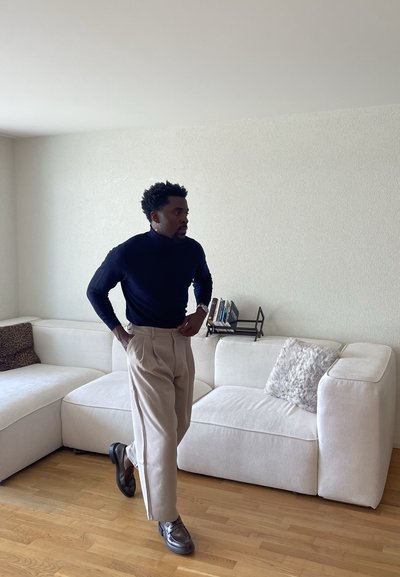 Hombre con cuello de tortuga negro y pantalones beige de pie con las manos en los bolsillos en una sala de estar con un sofá seccional blanco y suelo de madera.