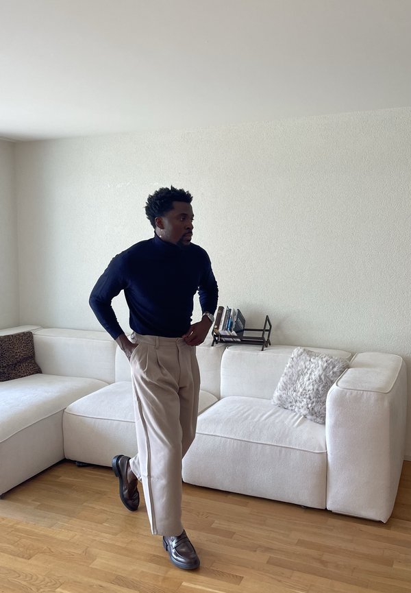 Mężczyzna w czarnym golfie i beżowych spodniach stoi z rękami w kieszeniach w salonie z białą sofą narożną i drewnianą podłogą.