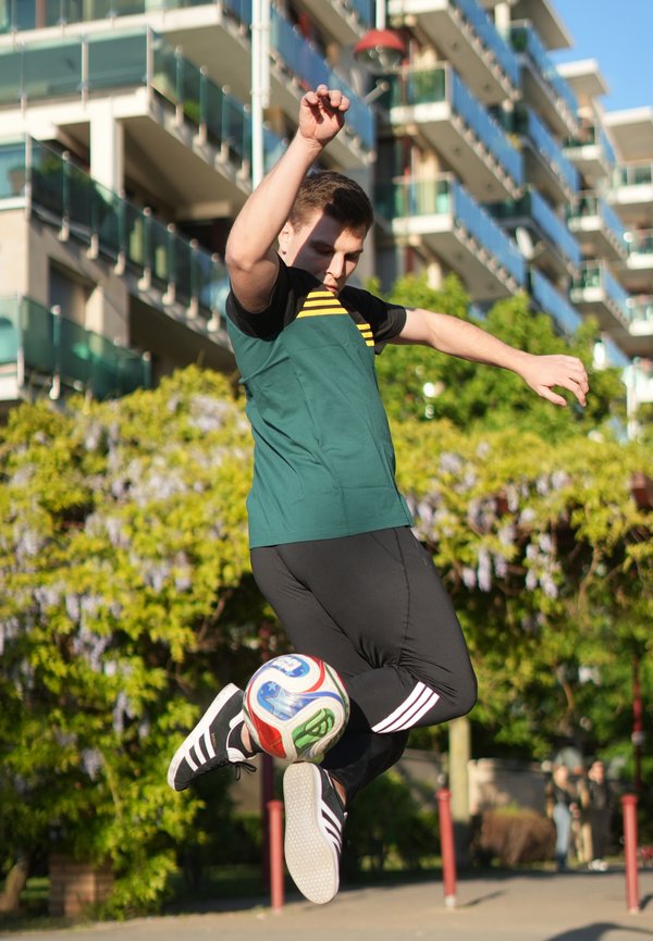 Homme en tenue de sport verte et noire sautant pour donner un coup de pied à un ballon de football en plein air, près de bâtiments modernes et d’arbres, sous le soleil.