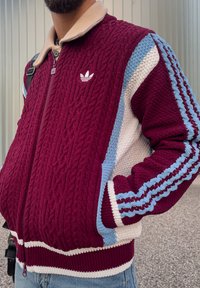 Vínový pletený sveter so zapínaním na zips, béžovým golierom a dizajnom farebných blokov v modrej, bielej a vínovej farbe s tromi pruhmi na rukávoch.